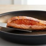A photorealistic image shows a thick-cut salmon fillet perfectly pan-seared with a golden-brown crust in a non-stick pan. A metal spatula slides easily underneath, revealing a visible opaque cooked band on the fish, emphasizing a flawless, easy sear.