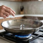 A photorealistic close-up of a single, spherical water droplet dancing on a superheated, brushed stainless steel frying pan, demonstrating the Leidenfrost effect. A cook's hands are visible in the foreground, intently watching the droplet. The background is a subtly blurred, minimalist kitchen counter with natural light.