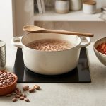 A photorealistic kitchen still life demonstrating the stages of cooking dried beans. Dry beans and whole tomatoes are in the foreground, followed by beans simmering without tomatoes, and finally, tender beans in a rich tomato sauce.