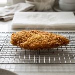 A photorealistic image shows a perfectly crisp, golden-brown pan-fried breaded cutlet (or similar food item) resting on a silver wire cooling rack on a clean, light kitchen counter. Its dry, textured breading appears shatteringly crispy, free from oil or moisture. Heat haze shimmers above, while steam rises from underneath the food through the rack, illustrating efficient cooling. In the soft-focus background, paper towels show a faint cloud of condensation above them, highlighting the rack's superior method for preventing soggy breading.