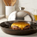 A pristine stainless steel dome covers a sizzling gourmet beef burger with glossy, melted cheddar cheese in a cast iron skillet. Subtle steam escapes from the dome on a clean kitchen counter, lit by soft natural light.