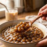 A close-up of glistening, dark brown tapioca pearls in warm, golden-brown syrup, gently stirred by hands with a wooden spoon, in a softly blurred, inviting kitchen setting.