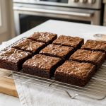 A close-up of perfectly baked brownies, featuring an impossibly glossy, tissue-thin crust with delicate cracks that reveal a rich, dark chocolate interior. They are precisely cut into uniform squares and neatly arranged on a minimalist surface in a clean, modern home kitchen, bathed in soft, natural light that highlights their exquisite sheen and delicious perfection.