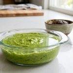 A photorealistic image shows a clear glass dish filled with vibrant, bright green mashed avocado, perfectly topped with a thin, reflective layer of water. In the soft-focus background, a ceramic bowl holds unappetizingly brown, oxidized avocado. A discarded lime wedge and crumpled plastic wrap are visible on a clean, brightly lit countertop.