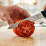 A hyper-realistic macro shot of a sharp serrated utility knife making a precise, clean cut into the firm, glossy skin of a vibrant red heirloom tomato. The blade's micro-teeth are visibly engaged, guided by a blurred chef's hand, on a light wooden cutting board with shallow depth of field, emphasizing effortless precision.