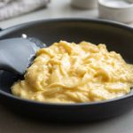 A close-up of perfectly cooked, creamy pale yellow scrambled eggs with delicate curds, glistening subtly in a dark non-stick skillet. A minimalist silicone spatula rests nearby. Bright, soft natural light illuminates the scene against a subtly blurred kitchen counter.