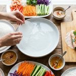 An overhead, photorealistic shot of a vibrant, meticulously clean summer roll assembly station. Hands gently prepare rolls amidst artfully arranged fresh julienned vegetables (carrots, cucumbers, bell peppers, mint, cilantro), dry rice paper wrappers, and small bowls of peanut and nuoc cham dipping sauces. A hydrated wrapper softens in a central water bowl, bathed in bright, natural light. The scene is perfectly organized and inviting.