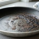 A close-up, stark image shows the interior of a nonstick frying pan with severe chemical damage. Dark, glossy, tar-like polymerized residue clings unevenly to the surface, contrasting sharply with the dull, degraded original nonstick coating. In the blurred background, a generic cooking spray can is angled towards the pan, implying its role in the damage. The scene is brightly lit on a clean kitchen counter.