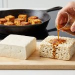 A photorealistic close-up of tofu transformation in a clean, brightly lit kitchen. On a wooden board, a smooth, fresh white tofu block sits beside a highly porous, sponge-like block. A hand pours amber marinade onto the porous tofu, which instantly absorbs it. In the soft-focus background, golden-brown, pan-fried tofu pieces rest in a cast iron skillet.