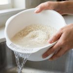 A close-up shows hands gently washing white short-grain rice in a sleek, modern Japanese rice washing bowl. Cloudy water swirls around the rice inside the bowl, while clear water cascades from side drainage slots into a blurred kitchen sink. The naturally lit scene conveys calm efficiency and cleanliness.