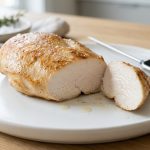 A close-up, photorealistic shot of a perfectly cooked, succulent golden-brown chicken breast on a white plate. A cut section reveals its moist, glistening white interior, with natural juices pooling on the plate. A silver digital meat thermometer rests beside it.