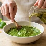 Photorealistic close-up of clean hands gently stirring vibrant green homemade pesto in a white ceramic bowl with a wooden spoon. A stream of golden olive oil pours into the smooth, glossy pesto, creating ripples. Fresh basil, pine nuts, garlic, and Parmesan cheese are softly blurred in the background, all bathed in bright natural light.