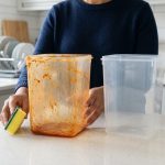 A photorealistic image of two clear plastic food containers on a modern kitchen counter. One is deeply stained with vibrant, greasy orange tomato sauce, while the other is sparkling clean. A pair of hands holds the stained container and a cleaning sponge, capturing a common domestic cleaning challenge.