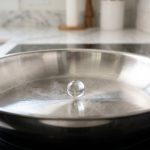 A photorealistic image of a gleaming stainless steel frying pan demonstrating non-stick cooking, either with a dancing water droplet or a perfectly sliding fried egg.