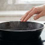A close-up, photorealistic image of a perfectly seasoned, deep black cast iron skillet with a satiny sheen, resting on a modern induction stovetop. Delicate wisps of steam rise from its surface as a human hand gently hovers above, checking for warmth or dryness in a pristine, minimalist kitchen.
