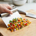 A human hand expertly scoops a vibrant mound of finely chopped vegetables from a clean wooden cutting board using a modern stainless steel bench scraper. A gleaming chef's knife rests in the soft-focus background, conveying kitchen efficiency and cleanliness.