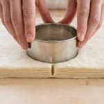 Photorealistic close-up of hands precisely pressing a biscuit cutter into raw, laminated dough, revealing distinct layers and a clean, unsealed vertical cut.