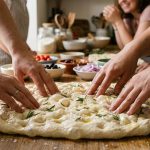 Close-up of diverse hands gently dimpling soft, pliable focaccia dough with deep indentations. Glistening olive oil, coarse sea salt, and fresh rosemary adorn the dough on a warm wooden counter bathed in soft natural light. In the softly blurred background, a cozy kitchen with focaccia toppings and hints of other people suggests communal baking.