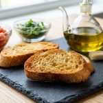 A perfectly toasted golden crostini in sharp focus, alongside more toasted baguette slices on a minimalist platter, an olive oil cruet, and a pastry brush. The blurred background shows bowls of fresh diced tomatoes and green basil, hinting at a DIY bruschetta station. Bathed in bright natural light.