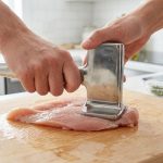 Close-up of clean hands using a flat, polished stainless steel meat pounder to evenly flatten a raw chicken breast on a light wooden cutting board. A subtly blurred, clean modern kitchen is in the background.
