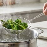 A gender-neutral hand expertly scoops vibrant green blanched broccoli and asparagus from a steaming pot of boiling water using a wire spider skimmer, with water cleanly draining. A blurred modern kitchen counter is in the background.