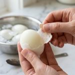A close-up, photorealistic shot of a hand gently peeling a hard-boiled egg. The shell separates cleanly, revealing a smooth, pristine egg white with a visible membrane. Steam rises subtly from the warm egg. In the soft-focus background, an ice bath with other eggs hints at the cooling process in a bright kitchen.