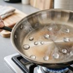 A pristine, preheated stainless steel skillet in a photorealistic close-up. Several perfectly spherical water droplets shimmer and dance on a visible cushion of steam, demonstrating the Leidenfrost effect. A hand gently tilts the pan in a softly blurred, minimalist kitchen background.