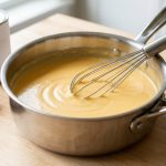 A perfectly smooth, velvety golden cheddar cheese sauce shimmers in a gleaming stainless steel saucepan, with a whisk gently resting in it. It sits on a clean kitchen counter, with a blurred background showing an evaporated milk can, shredded cheddar, and macaroni pasta, all bathed in soft, warm light.