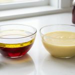 A pristine, well-lit modern kitchen counter displays two clear glass bowls in a photorealistic food photography style. The left bowl shows distinctly separated golden olive oil and red wine vinegar. The right bowl holds a creamy, homogenous pale yellow emulsified vinaigrette, with mustard, honey, and a whisk nearby, illustrating the science of emulsions.