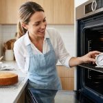 A person in a modern, sunlit kitchen stands before an open oven, intently looking at a gleaming stainless steel oven thermometer inside. A perfectly golden-brown cake sits nearby, a testament to precise baking. The scene is clean, photorealistic, and highlights accurate temperature control.