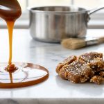 A photorealistic close-up contrasting two types of caramel on a white marble counter: on the left, smooth, glossy amber caramel drips and pools; on the right, dull, gritty, crystallized caramel with visible sugar crystals. A blurred background shows a saucepan and corn syrup.