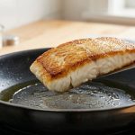 A close-up, photorealistic image of an appetizing, perfectly seared, golden-brown white fish fillet being lifted cleanly from a gleaming non-stick pan with a metal spatula, against a blurred modern kitchen background.
