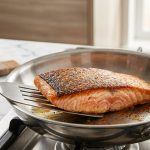 **Alt text:** Close-up of a salmon fillet with perfectly crispy, golden-brown skin being lifted from a stainless steel skillet with a metal spatula. A jar of mayonnaise and a pastry brush are visible on a marble countertop in the soft-focus kitchen background.