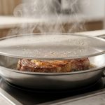**Alt text:** A thick ribeye steak searing in a stainless steel skillet on an induction stove, covered by a fine-mesh splatter guard with steam rising through it.