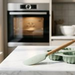 **Alt Text:** A minimalist photograph of a pastel-colored silicone spatula and matching baking mold with fresh water droplets, resting on a light marble countertop. Bright, natural morning light illuminates the scene, with a softly blurred modern kitchen and glowing oven in the background.