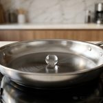 **Alt text:** A macro photograph of a single, perfect sphere of water gliding across a hot, polished stainless steel skillet, demonstrating the Leidenfrost effect in a bright, modern kitchen.