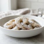 **Alt text:** Macro photograph of fresh, raw, peeled shrimp in a white ceramic bowl on a marble countertop, glistening under natural light and sprinkled with coarse sea salt.