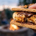 **Alt text:** Close-up of a gourmet s'more with a toasted, gooey marshmallow and melting dark chocolate between graham crackers, set against a blurred background of a warm backyard fire pit and glowing string lights.