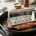 **Alt Text:** Close-up of a rectangular, vented stainless steel chef's press resting on a sizzling steak in a cast-iron skillet, with steam rising from the vents and a golden-brown crust forming on the meat.