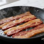 **Alt Text:** A close-up photograph of flat, thick-cut bacon cooking in a dark skillet with simmering water and gentle steam, set against a blurred kitchen countertop.