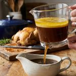 **Alt Text:** A close-up photo of a glass fat separator on a rustic wooden countertop. Inside, a layer of glowing golden fat floats cleanly atop rich, dark beef broth, which pours from the bottom release valve into a ceramic gravy boat. The background is a warm, soft-focus kitchen.