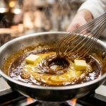 A macro close-up of a wire whisk stirring cubes of melting butter into a rich, glossy dark reduction sauce in a stainless steel skillet.