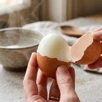 A close-up photograph of a hard-boiled brown egg being cleanly peeled on a gray stone countertop. Large, intact pieces of shell slide off to reveal a glossy, perfectly smooth white egg under bright, natural morning light.