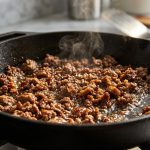 **Alt text:** A close-up shot of ground beef searing in a hot cast-iron skillet, showing a rich, mahogany caramelized crust, sizzling oil, and a light wisp of steam.