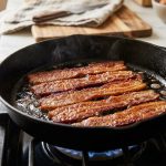 **Alt Text:** Thick-cut bacon strips sizzling in a cast-iron skillet, illuminated by warm morning light with a softly blurred kitchen background.