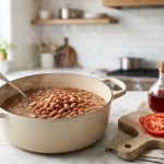 **Alt text:** A steaming cream-colored pot of simmering brown beans on a marble countertop, next to a wooden cutting board with a sliced red tomato, red wine vinegar, and a glass of red wine in a bright kitchen.
