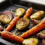 **Alt text:** A close-up of roasted vegetables—including caramelized Brussels sprouts, baby carrots, and red onion—with crispy, blistered edges on a dark baking sheet, sprinkled with sea salt and fresh rosemary.