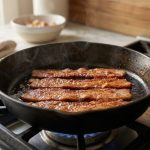 **Alt text:** A close-up photograph of thick, straight bacon strips cooking to a golden-brown crisp in a cast-iron skillet, glistening with rendered fat under soft, warm sunlight.