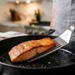 **Alt text:** Close-up of a flexible metal spatula sliding under a seared salmon fillet with a crispy, golden-brown crust in a cast-iron skillet.