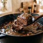 A close-up of rich beef stew simmering in a black cast-iron Dutch oven. A wooden spoon lifts a tender, glistening chunk of beef from the thick gravy, with a delicate wisp of steam rising in warm light.